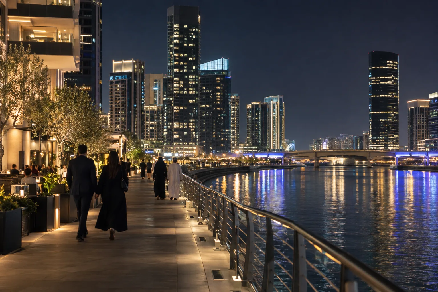 Al Maryah Island promenade at night, passers-by seen from behind in front of illuminated residential towers along the canal