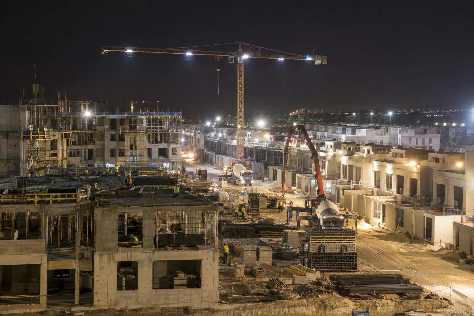 Suburban residential construction site lit at night, with rows of unfinished low-rise buildings and a crane