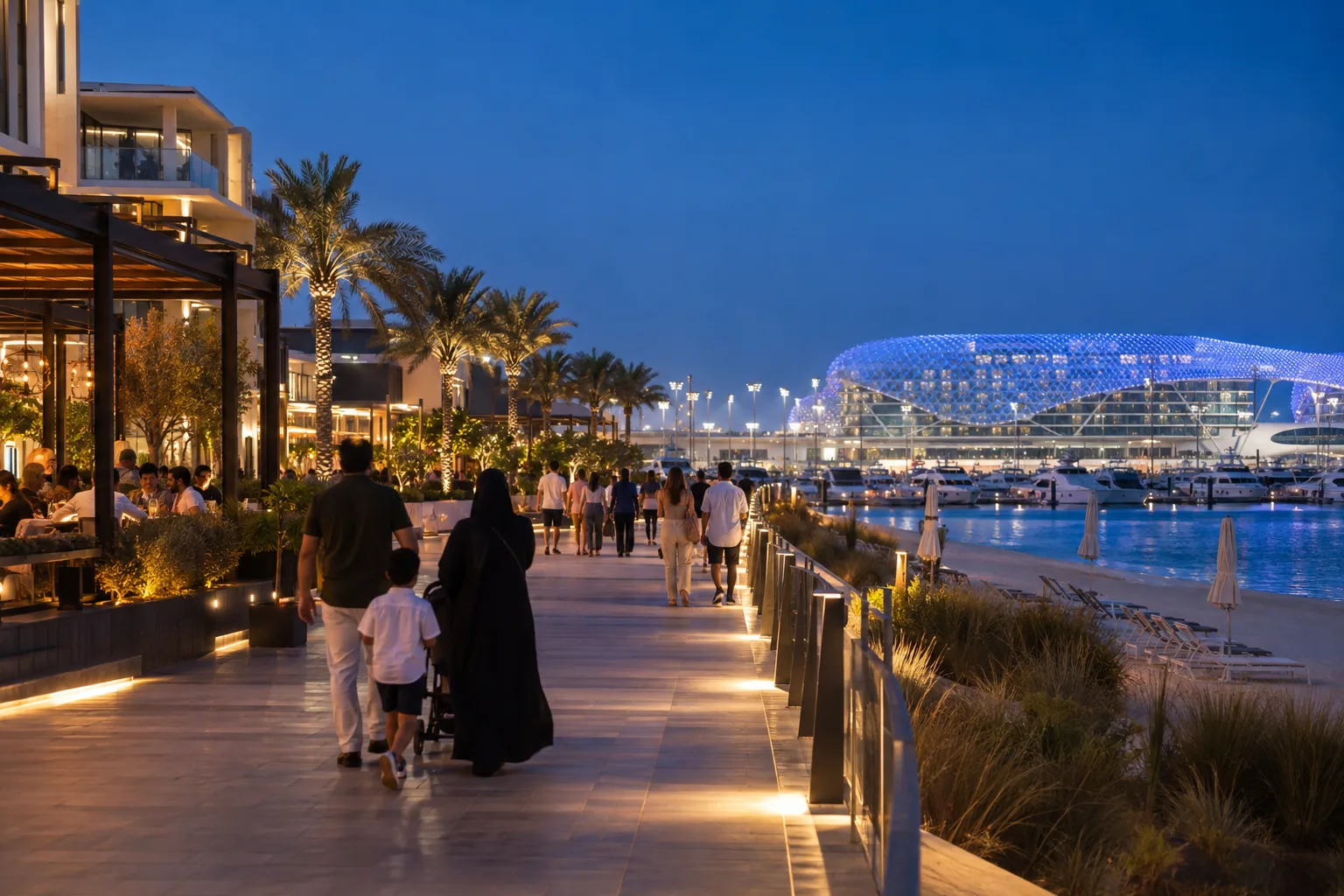 Yas Bay waterfront promenade at blue hour, strollers seen from behind, lit-up terraces and beach