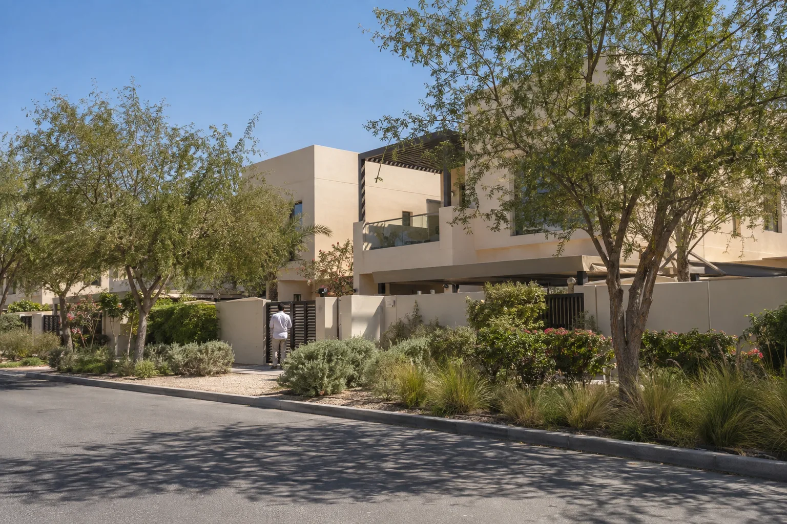 Sand-colored villas of a residential community in Abu Dhabi behind ghaf trees, an owner seen from behind at midday