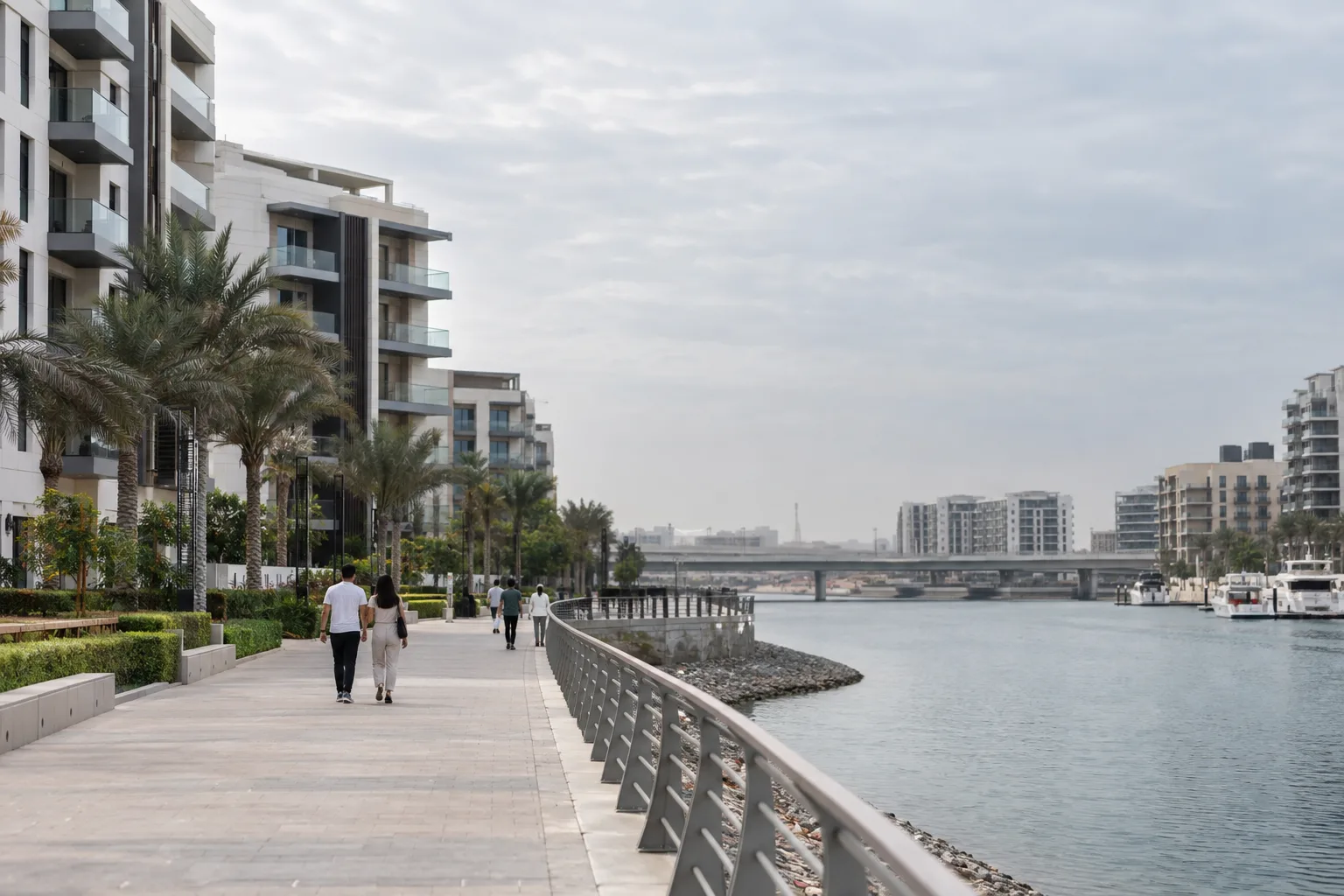 Walkers seen from behind along a canal-side promenade on Yas Island, beneath apartment buildings on an overcast day