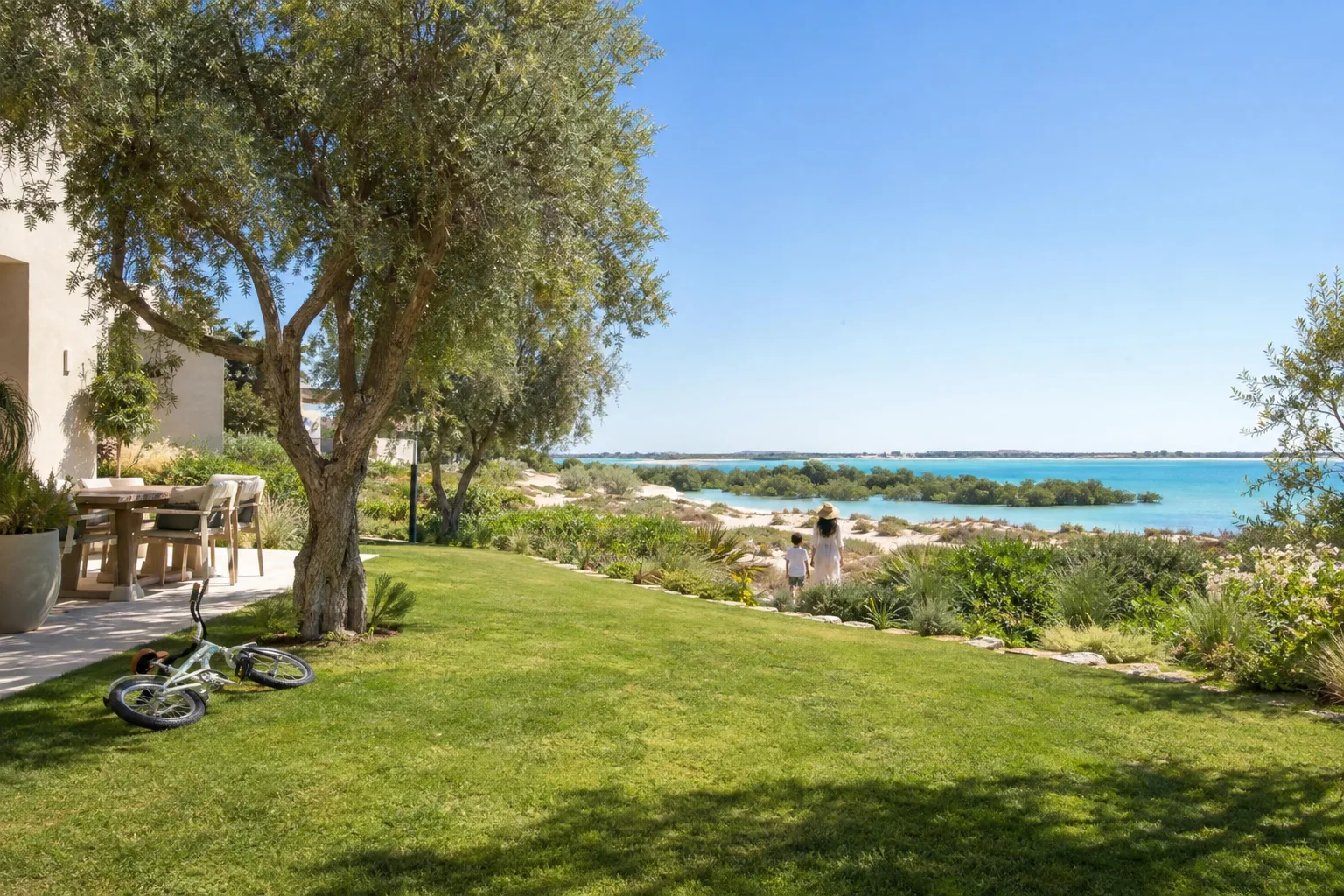 Villa garden in Al Jurf opening onto dunes and shallow sea, a child's bike lying on the grass